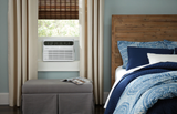 A Frigidaire FHWC064TE1 6,000 BTU Window Air Conditioner unit installed on a bedroom window, next to a bed.